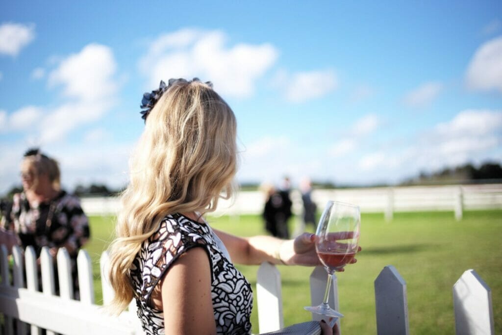 A woman looking away from the camera to the horse racing tack holding a glass of Koonara Wines on a bright sunny day.