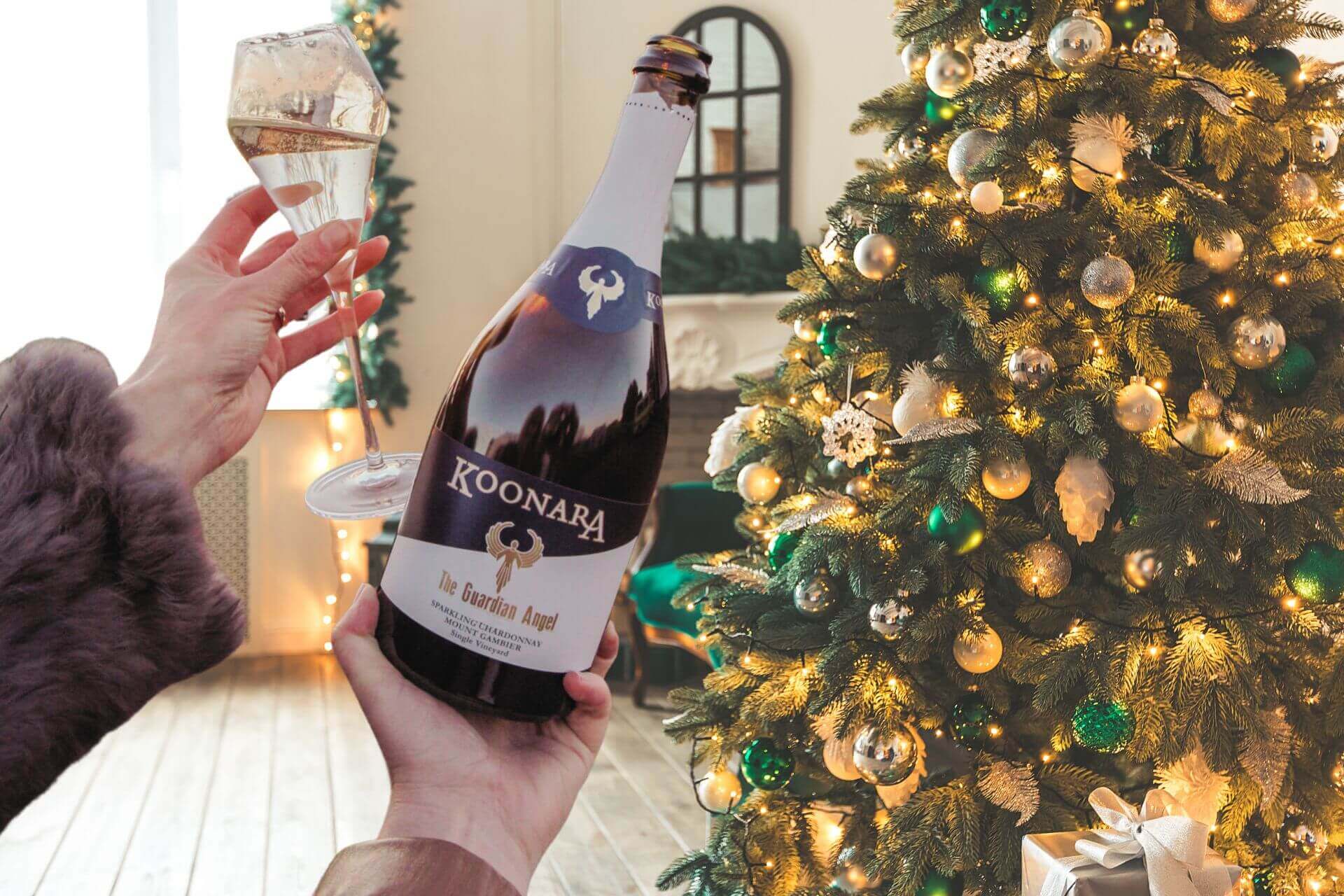 A woman is holding a bottle of Guardian Angel Sparkling Chardonnay by Koonara Wines in front of a Christmas tree.