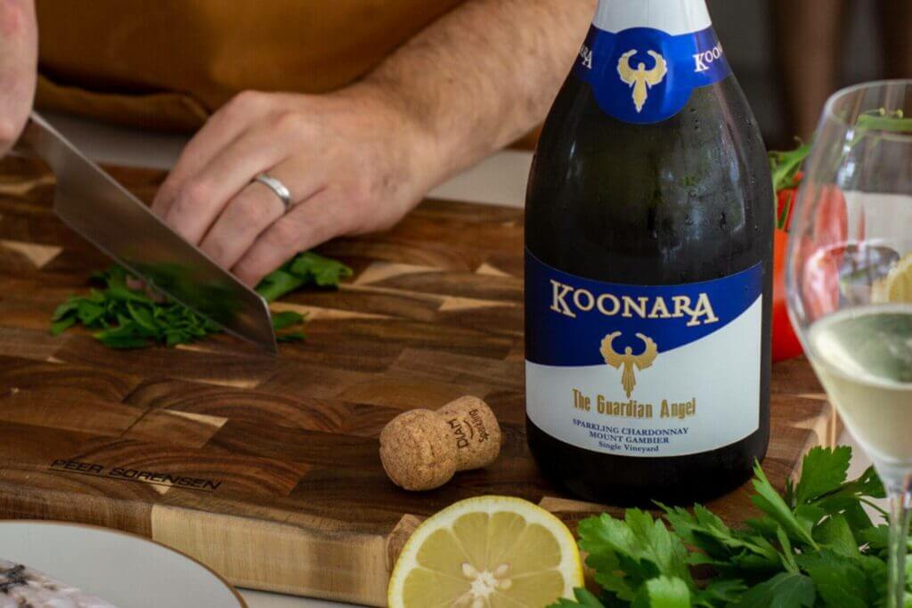 A man is slicing fish on a cutting board next to a bottle of Koonara Wines' Guardian Angel Sparkling Chardonnay.